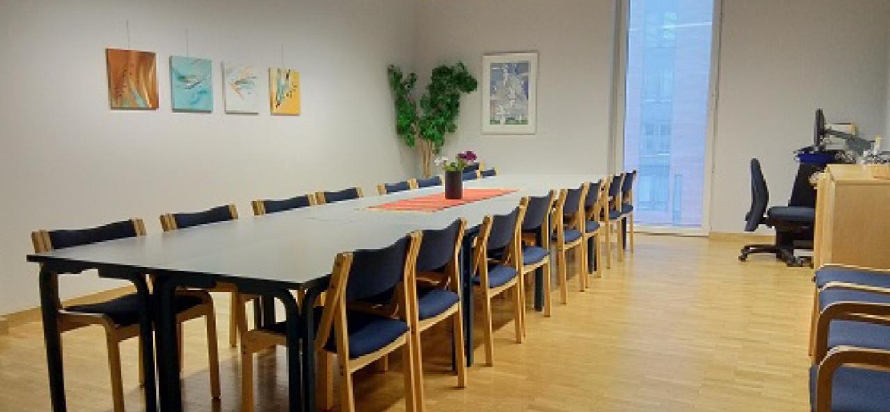 Pitkä pöytä, jonka ympärillä sinisiä tuoleja. Seinillä tauluja, nurkassa viherkasvi, keskellä pöytää kukkia. Takana yksi korkea ikkuna, ja huoneen nurkassa työpöytä ja -tuoli.